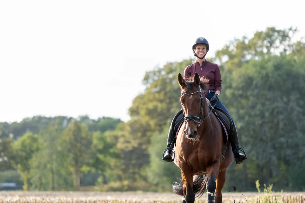 A front-facing shot of a rider in a burgundy shirt smiling while riding a chestnut horse toward the viewer in an open field.