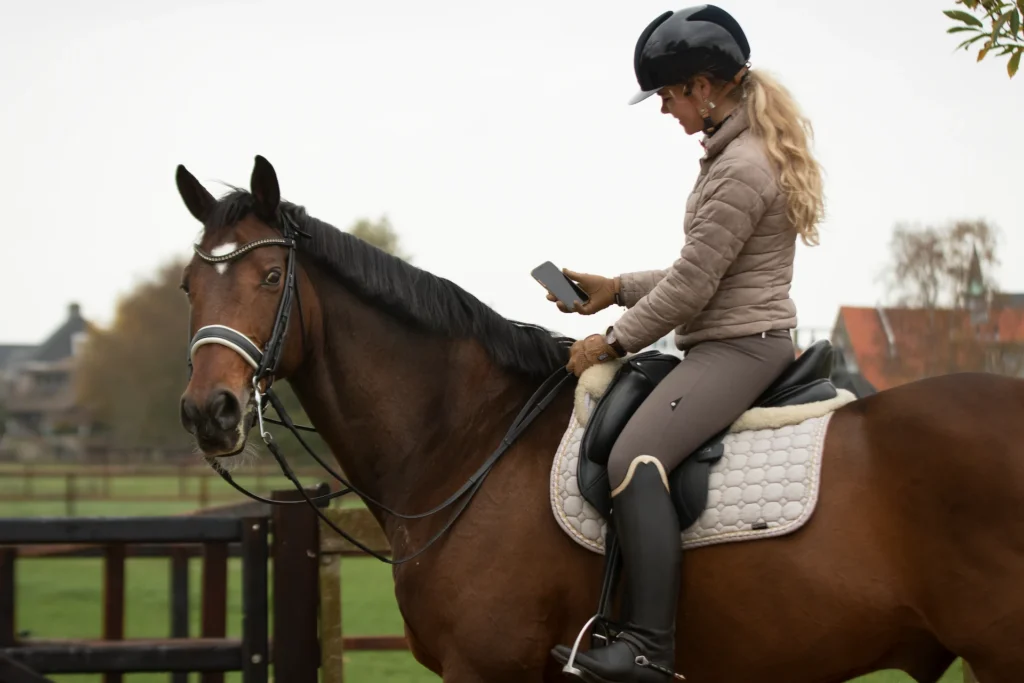 A side profile of a female rider sitting on a bay horse, holding a smartphone in her gloved hand to check data.