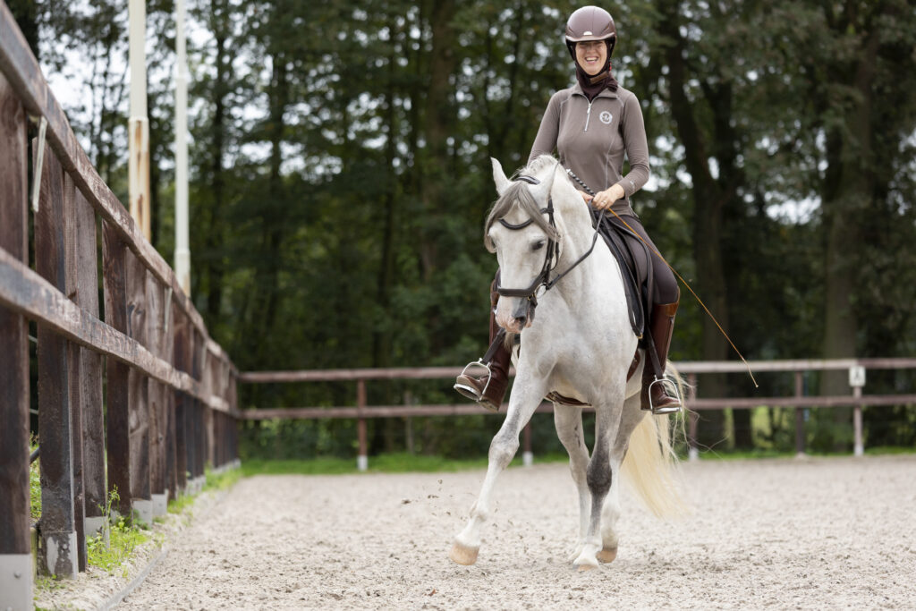 A smiling rider in a brown top and helmet rides a grey horse toward the camera in a sand arena.